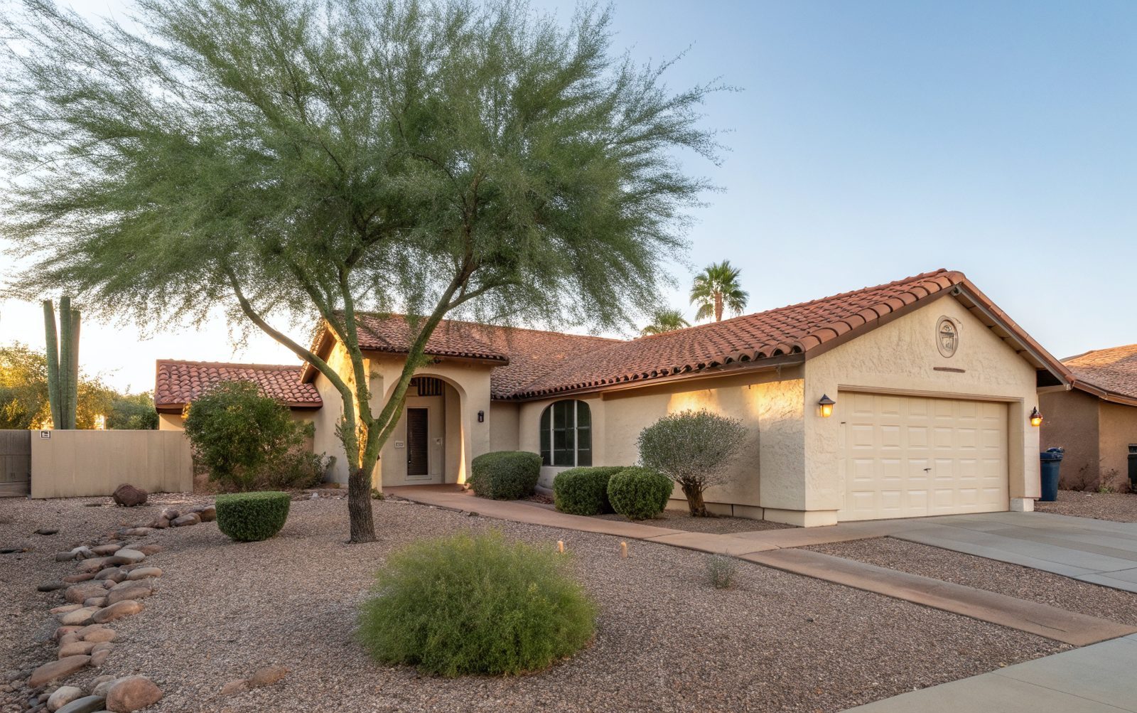 A one-story stucco home with a terracotta concrete tile roof, a palo verde tree and a gravel front yard