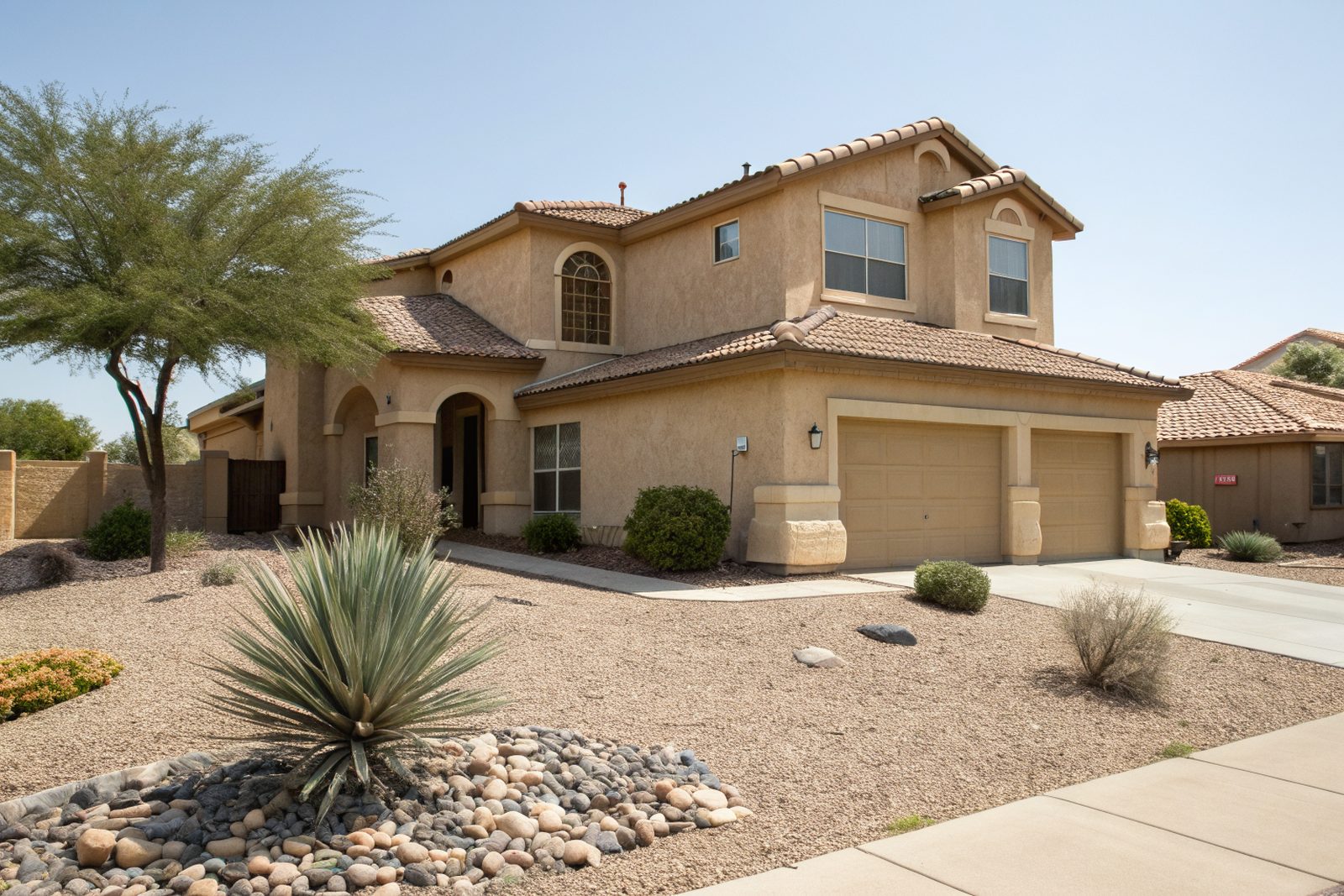 A two-story stucco home with a brown shingle roof and a desert-landscaped front yard